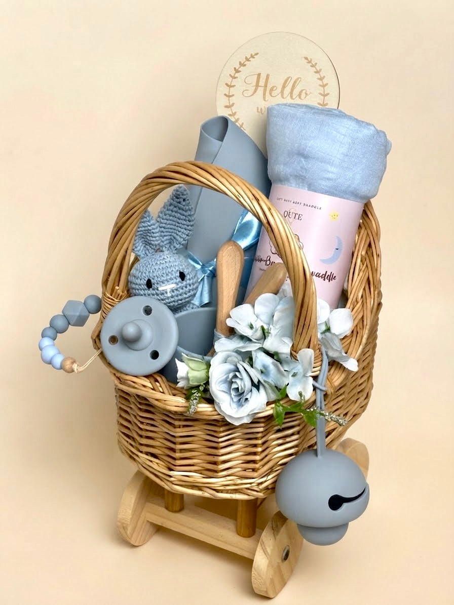 Luxury newborn baby boy feeding and blanket gift set in a wheeled wooden carriage basket by Qute Qatar, featuring a blue silicone feeding set, crochet toy, baby blanket, and floral accents for delivery in Doha.  