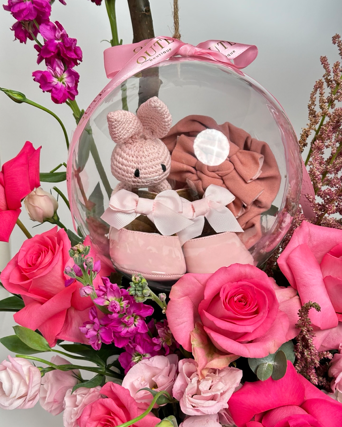 Lush white floral box filled with vibrant pink roses and purple stock, featuring a natural branch holding a clear bubble with a handmade crochet bunny and baby shoes.   