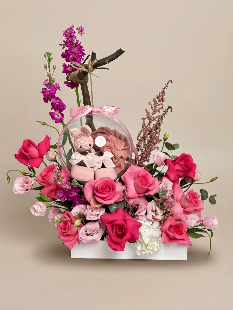 Baby Bloom Bubble newborn flower arrangement featuring pink roses, stock, and an acrylic keepsake bubble containing a mini crochet toy and baby booties.   