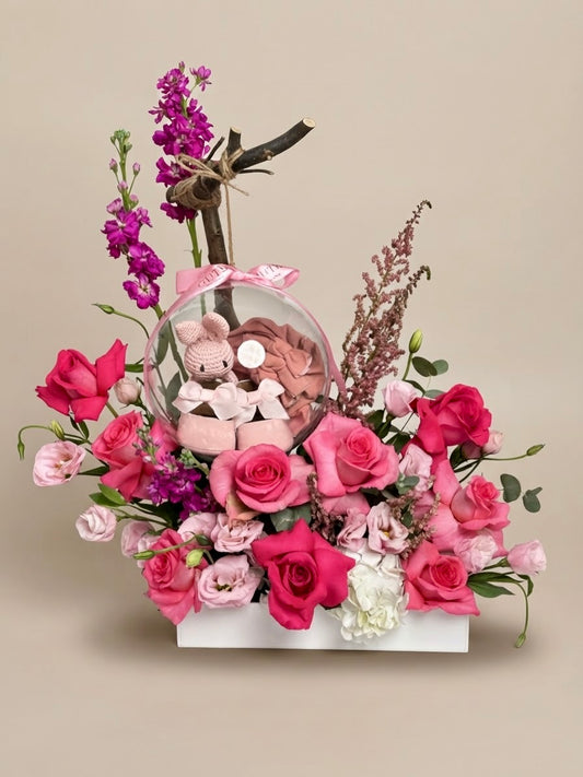 Baby Bloom Bubble newborn flower arrangement featuring pink roses, stock, and an acrylic keepsake bubble containing a mini crochet toy and baby booties.   