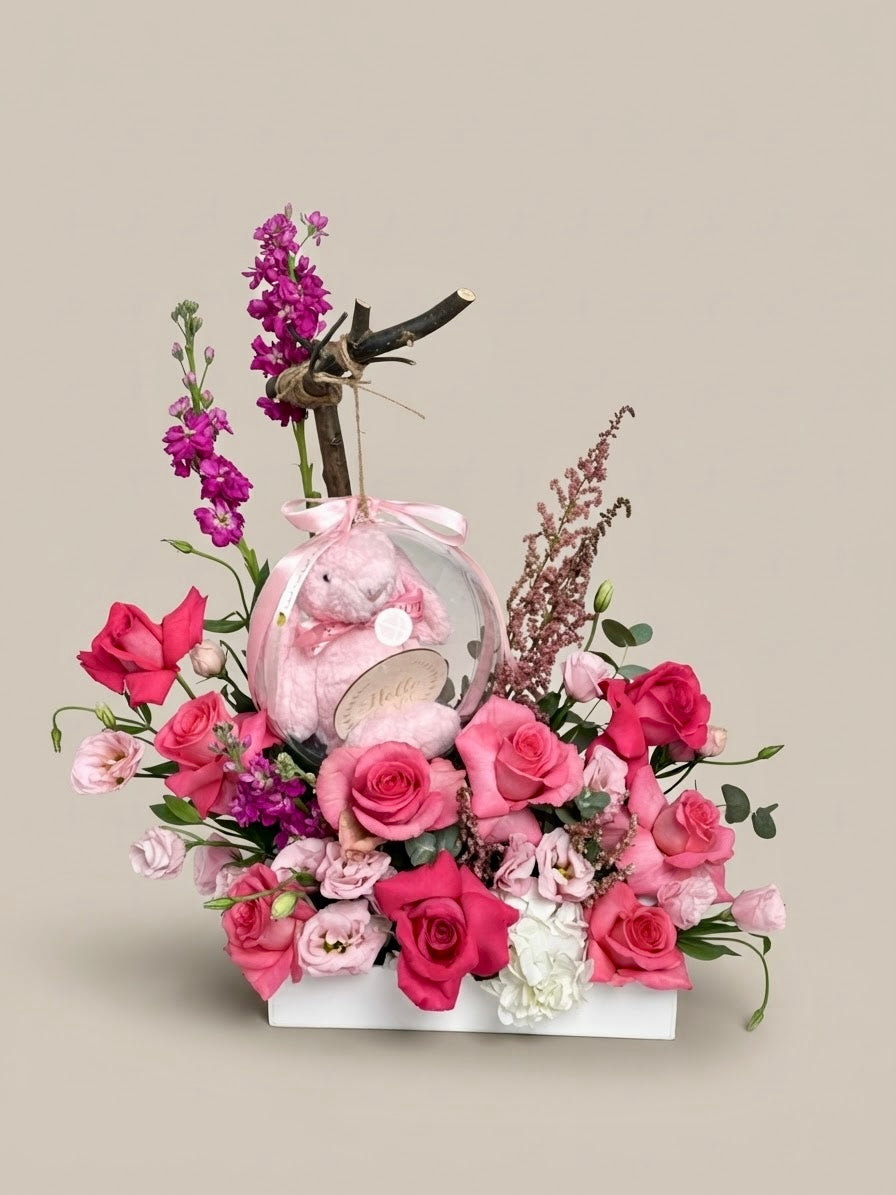 Fresh pink roses and stock flower arrangement in a white box featuring a clear acrylic keepsake bubble with a pink plush toy and "Hello Little One" tag, a Plaza Hollandi collaboration inspired by Seju from Ezdan Mall.   