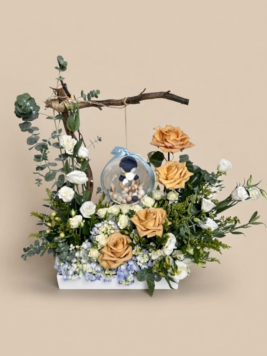 Baby Bloom Bubble newborn boy flower arrangement featuring champagne roses, blue hydrangeas, and an acrylic keepsake bubble with a crochet penguin toy and wooden teethers.