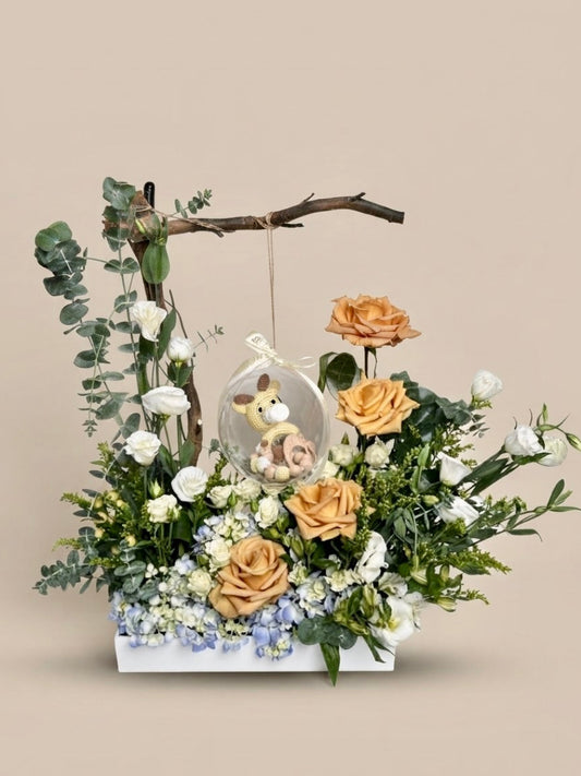 Baby Bloom Bubble newborn boy flower arrangement featuring champagne roses, blue hydrangeas, and an acrylic keepsake bubble with a crochet giraffe toy and wooden teethers.