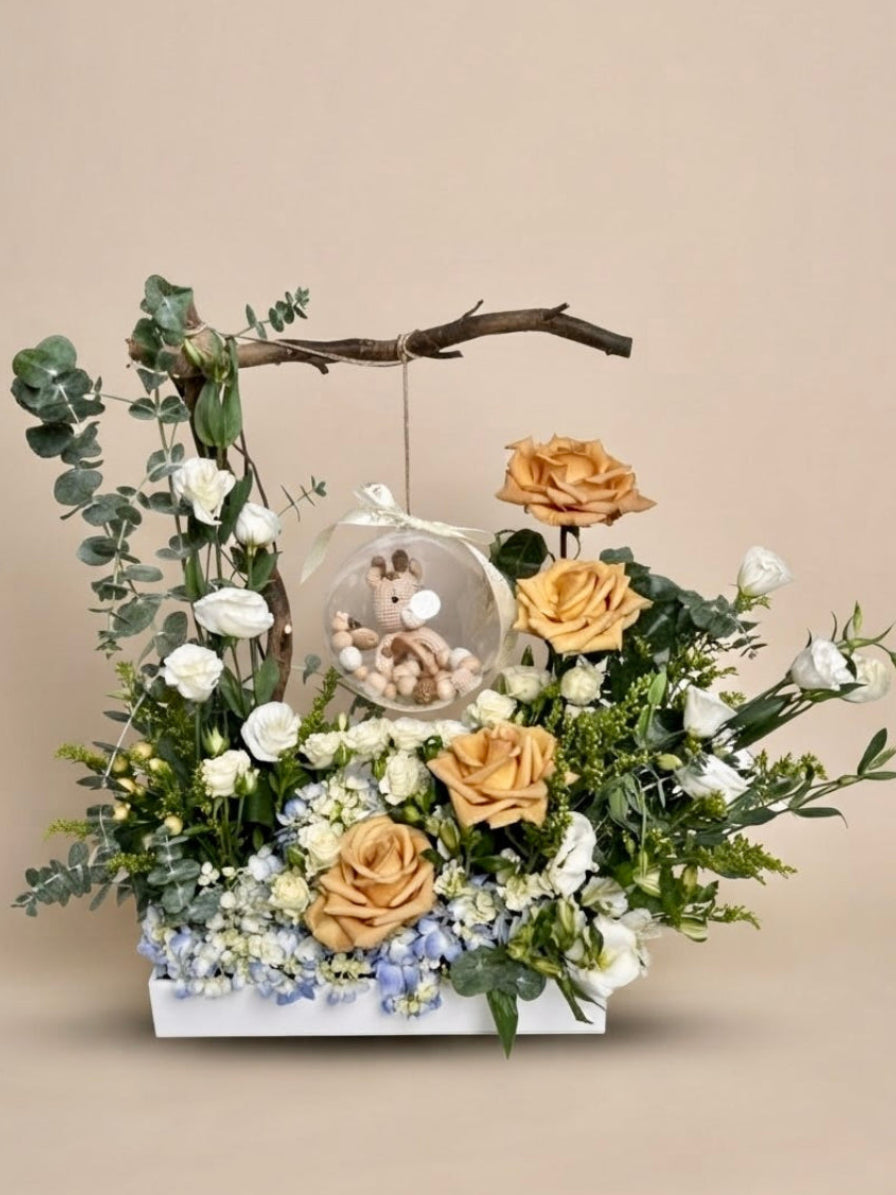 Baby Bloom Bubble newborn boy flower arrangement featuring champagne roses, blue hydrangeas, and an acrylic keepsake bubble with a crochet giraffe toy and wooden teethers.