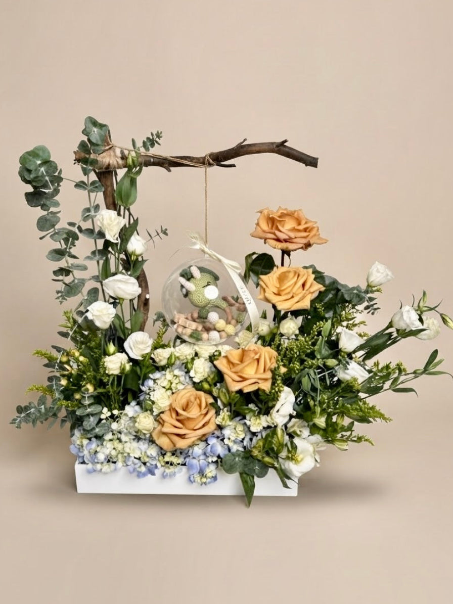 Baby Bloom Bubble newborn boy flower arrangement featuring champagne roses, blue hydrangeas, and an acrylic keepsake bubble with a crochet dinosaur toy and wooden teethers.   