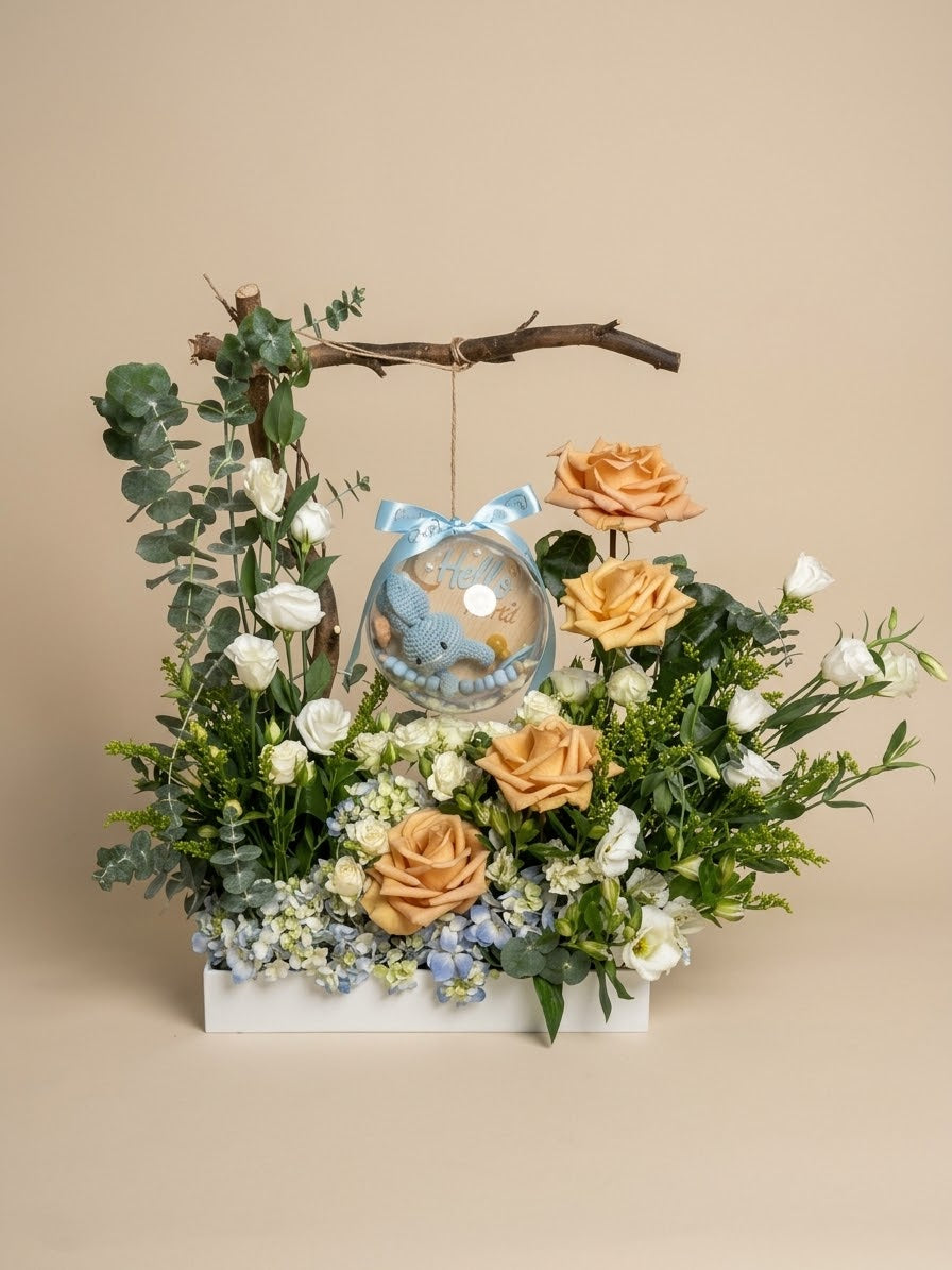 Baby Bloom Bubble newborn boy flower arrangement featuring champagne roses, blue hydrangeas, and an acrylic keepsake bubble with a crochet bunny rattle, pacifier, custom name clip, and Hello World card.