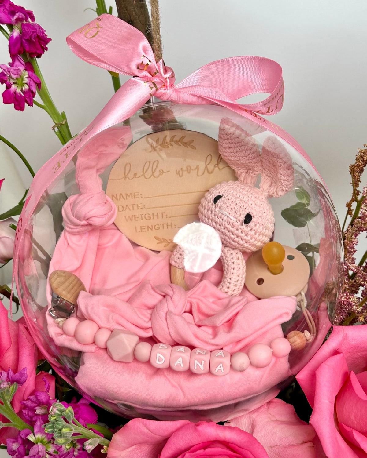 Lush white floral box filled with vibrant pink roses and purple stock, featuring a clear bubble holding a custom jersey swaddle, matching accessories, a pacifier, and a bunny toy.   