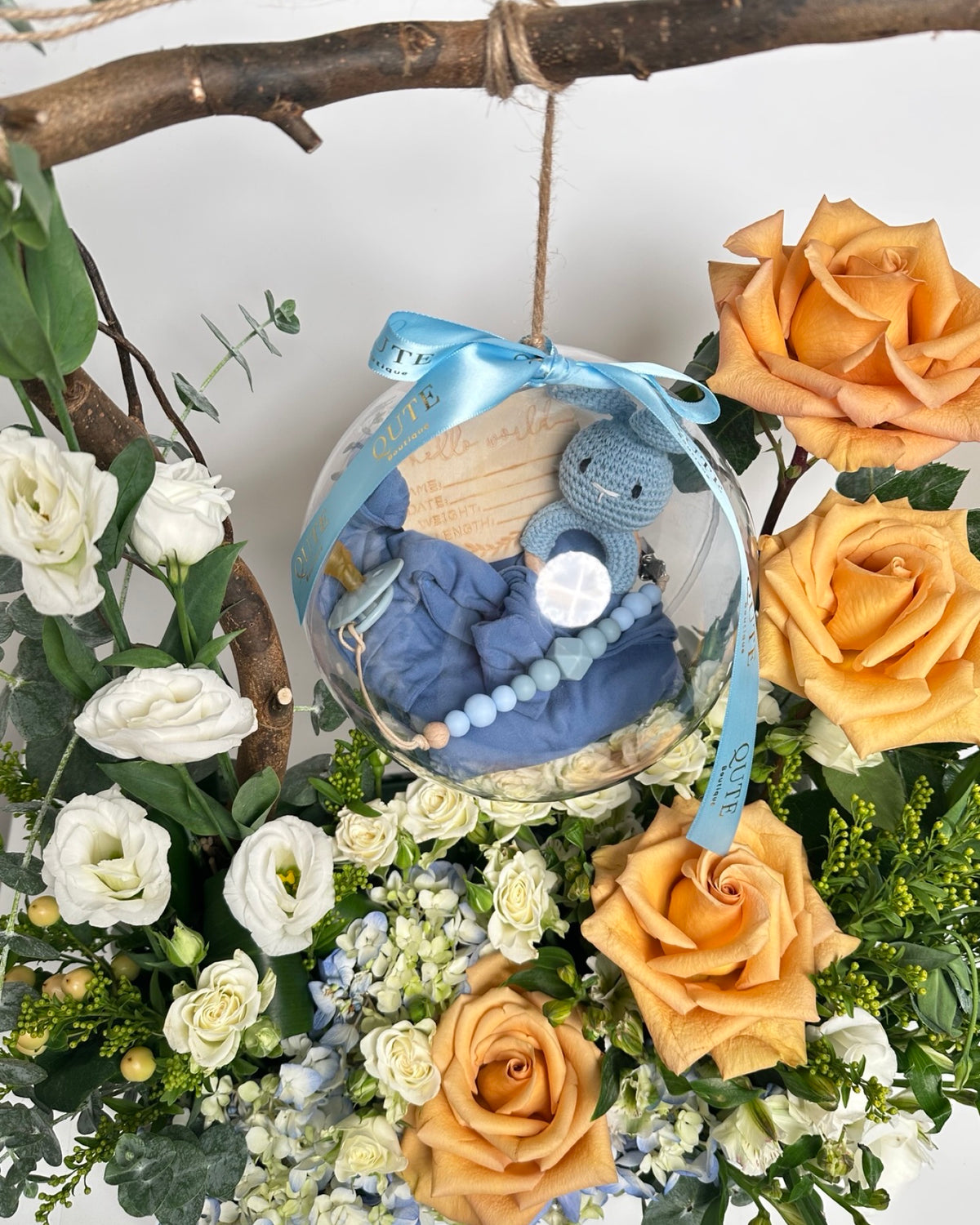 Lush white floral box filled with caramel roses, white eustoma, and blue hydrangeas, featuring a natural branch holding a clear bubble with a handmade crochet bunny and announcement tag.