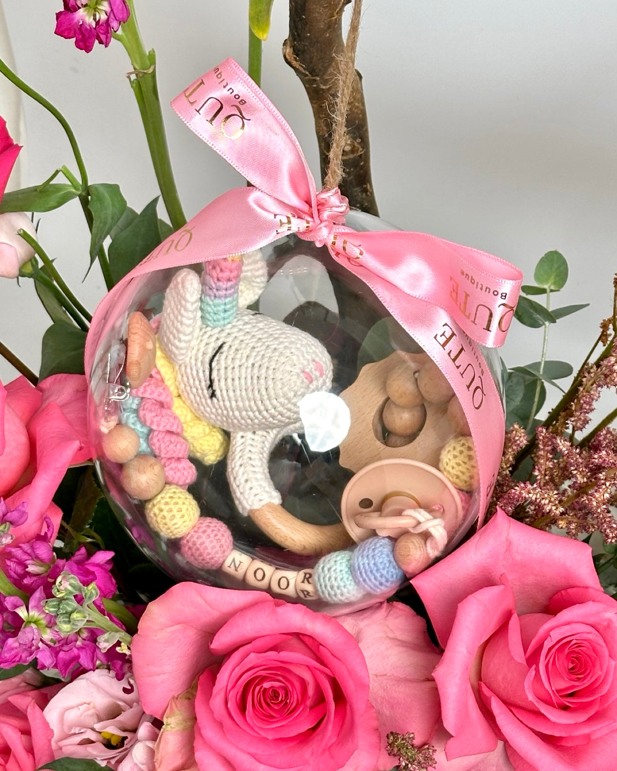 Lush white floral box filled with vibrant pink roses and purple stock, featuring a clear bubble holding a handmade unicorn toy, pacifier, teething ring, and personalized accessories.   
