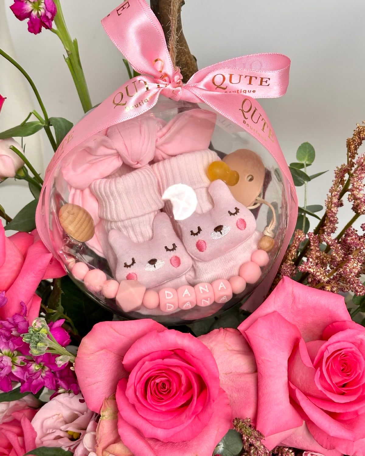 Pink rose and stock flower box arrangement with a hanging acrylic keepsake bubble enclosing a bow hat, booties, and a personalized name clip, styled by Plaza Hollandi at Ezdan Mall.   